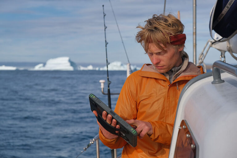 Sailor from the North West Expedition holding a 10-inch SailProof tablet while sailing, with glaciers in the background.
