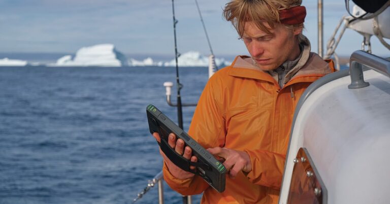 Sailor from the North West Expedition holding a 10-inch SailProof tablet while sailing, with glaciers in the background.