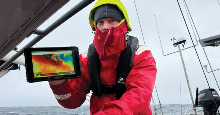 Sailor holding a SailProof tablet in the Northwest Passage, showing weather data with bright, clear lighting.