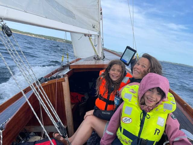 Una mujer y dos niños sentados en la cubierta de un velero clásico, uno de ellos sosteniendo una tableta SailProof.