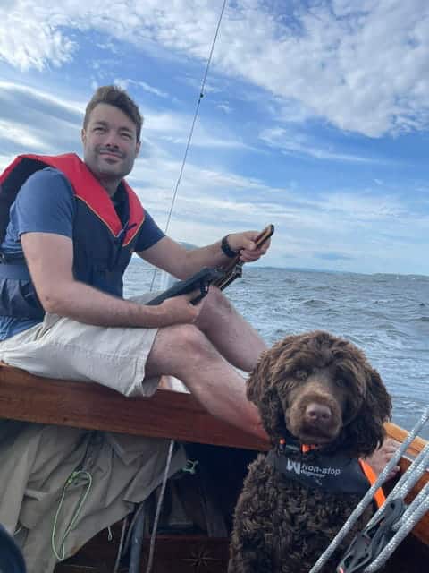Børre junto al timón de un velero clásico de madera, sosteniendo una tableta SailProof y con su perro a su lado.